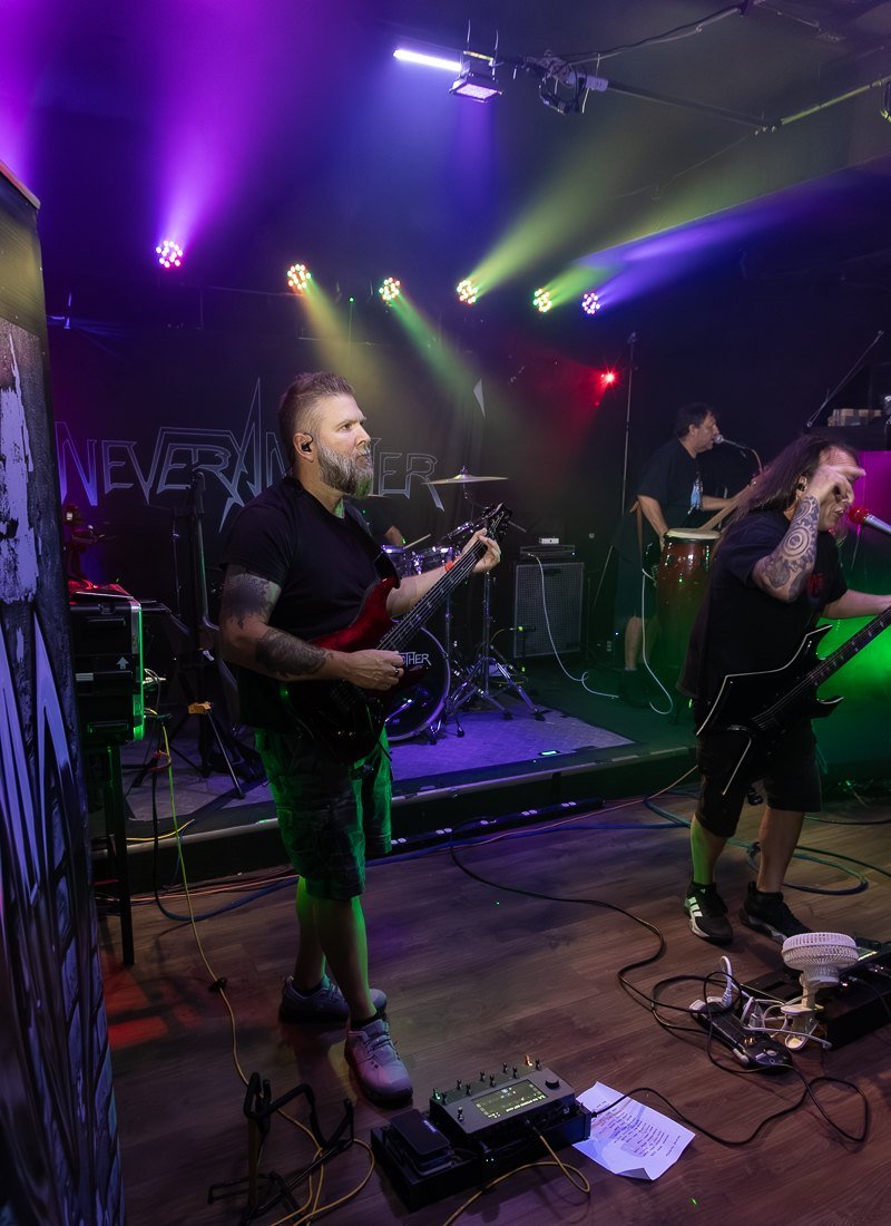 NEVERANOTHER mid-set under the stage rig, the band&rsquo;s backdrop banner lit behind them