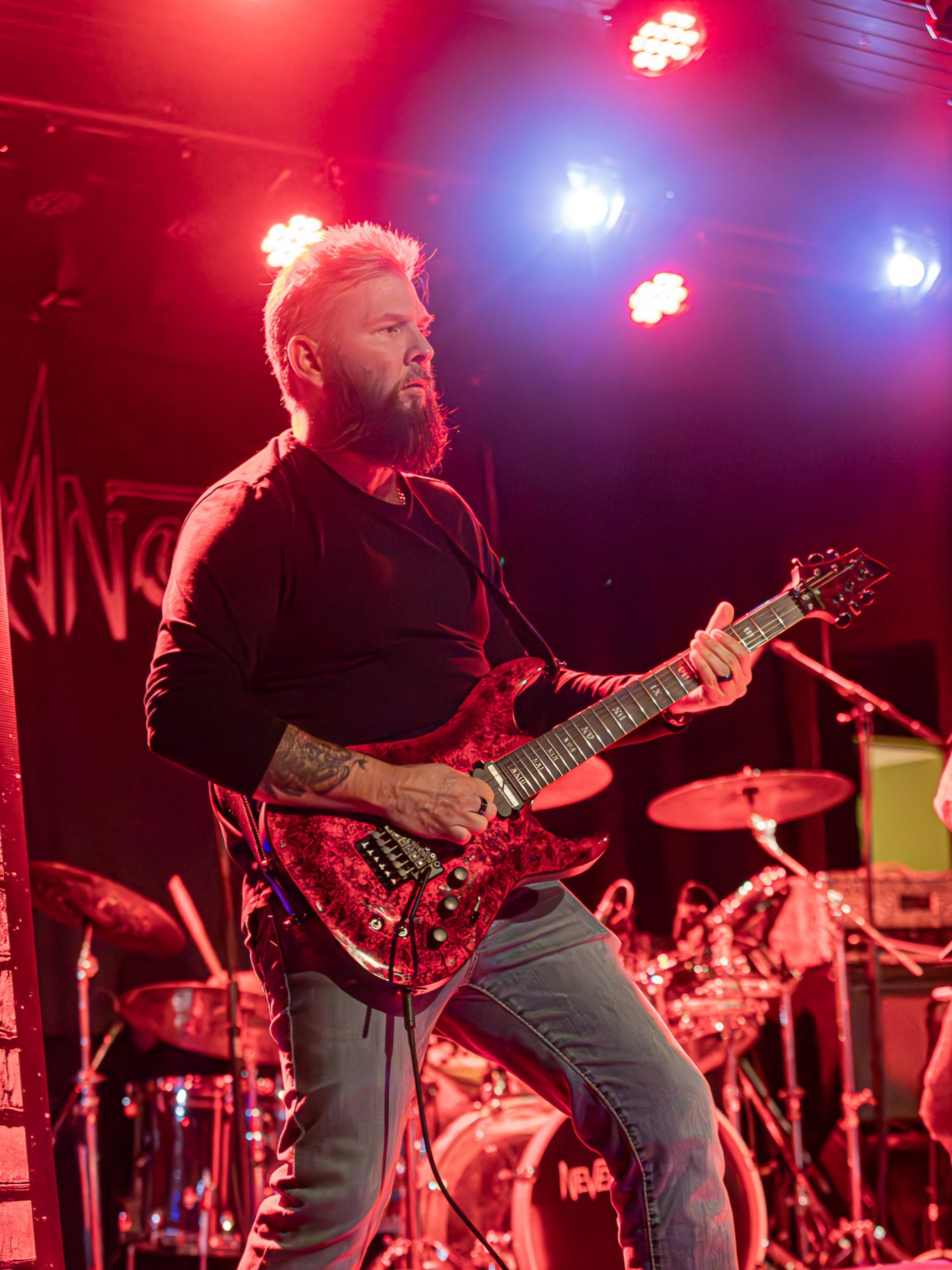 Vic Gratwicke mid-riff under red and blue stage lights, the NEVERANOTHER banner behind him