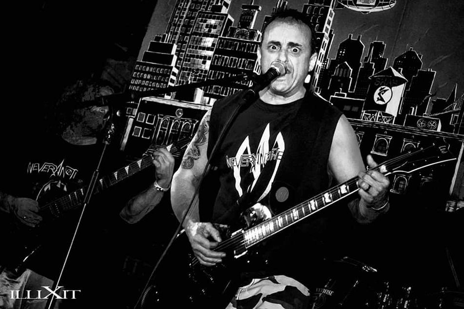 Black and white: the band mid-song in NEVERANOTHER shirts against a painted city-skyline backdrop